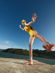 Obraz premium Yellow swimsuit woman standing on rocky edge near water and mountains in background, capturing a scenic and adventurous moment by a natural landscape