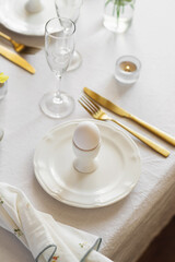 Happy Easter! Spring elegant table setting with white plates, egg cups, gold cutlery and vases with yellow flowers on a white tablecloth
