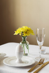 Happy Easter! Spring elegant table setting with white plates, egg cups, gold cutlery and vases with yellow flowers on a white tablecloth