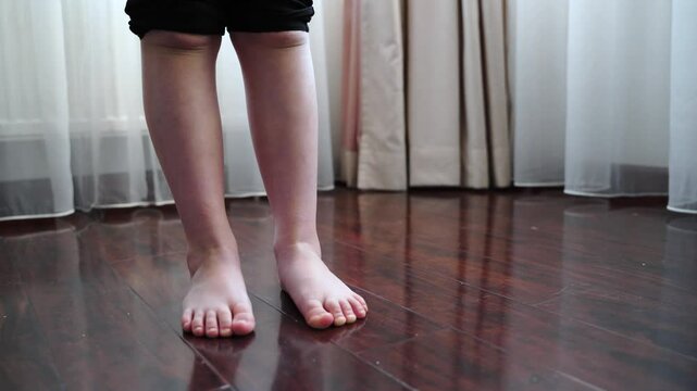Girl stands barefoot on a wooden floor. Legs are visible up to the knees. She shifts weight from foot to foot. Heels lift alternately. Black shorts cover upper legs. Balance and movement focus