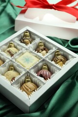 Box with tasty sweets in shape of baubles on green fabric background, closeup
