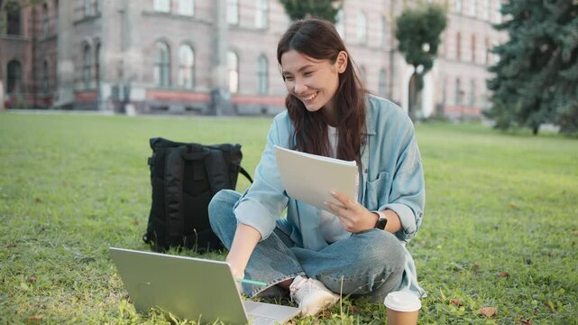 Charming Caucasian girl sitting on grass at college campus. Holding notepad with one hand. Typing on keyboard. Searching for educational video or answer online. Backpack laying on ground.