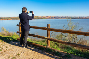 Man taking pictures with mobile phone of the landscape formed by the mountains and Lake Castrejon, Toledo, Spain.