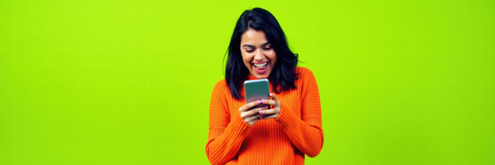 Fototapeta premium A joyful woman in an orange sweater engages with her smartphone against a vibrant green backdrop, capturing the essence of digital communication and modern lifestyle.