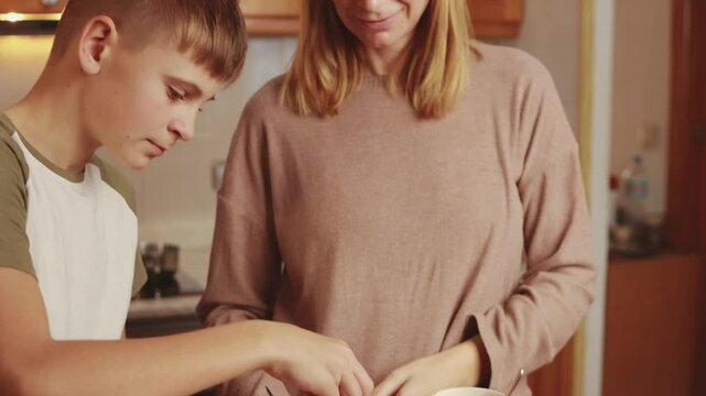 Caring mom sprinkling marshmallows into steaming hot chocolate, bonding with teenage son in sunlit kitchen during peaceful morning moments