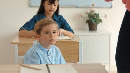 Bored elementary student looking at teacher while attend in classroom. Diverse smart student sitting at classroom while listening teacher explain about classwork or test. Education concept. Pedagogy.