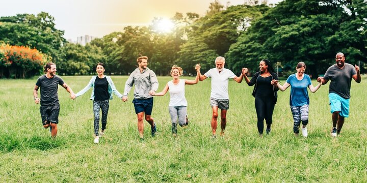 Group of diverse people holding hands, running in a park. Smiling men and women enjoying outdoor activity. Unity in nature. Diverse group, joyful moment. Diverse people group in park outdoor. - Powered by Adobe