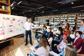 Diverse group of students in a library listens to teacher presenting at a whiteboard. Diverse...