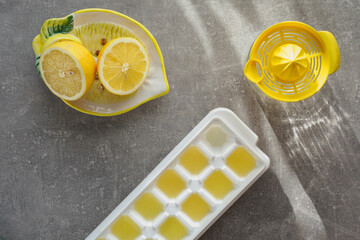 Halved lemon, yellow citrus juicer, white ice cube tray with fresh lemon juice. Overhead shot.