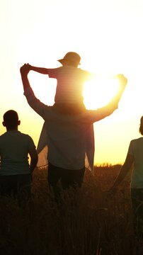 Dark silhouettes of happy family on yellow sunset in natural field. Father mother parents children kids walk along field, heading for golden sun, holding hands. Concept of bonding, strong family ties.