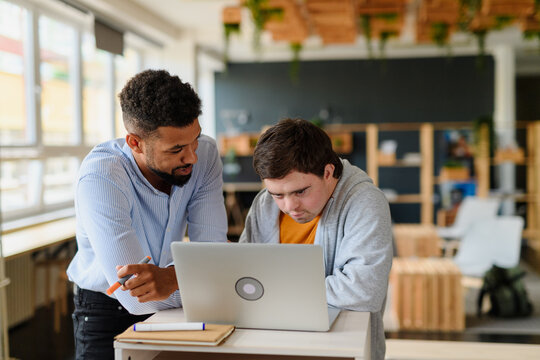 Happy young man with Down syndrome and his tutor using lapotp indoors at school.