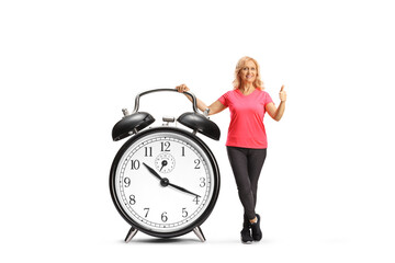 Mature woman in sportswear posing with a big alarm clock and gesturing thumbs up
