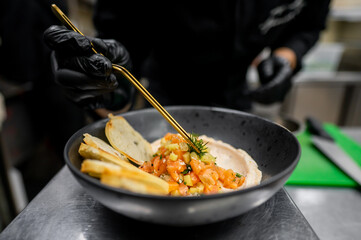 A chef in a black glove carefully plates a vibrant dish featuring fresh vegetables and a creamy base, accompanied by crispy toasted bread, showcasing culinary artistry in a modern kitchen.