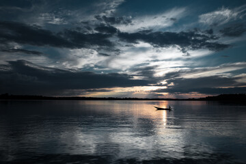 A lone boat floats on a tranquil lake, silhouetted against a moody sky with golden sunset reflections on the water. The vast horizon and cloud formations create an atmospheric and cinematic scene.