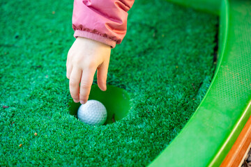 Child Hand Retrieving Golf Ball from Hole on Mini-Golf Course. Little girl playing mini golf outdoor. Family vacations or resort.