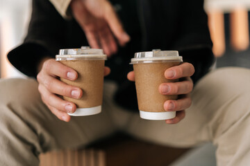 Cropped shot of unrecognizable friendly man holding two disposable coffee cups, conveying warm gesture of sharing beverage in cozy, relaxed setting, perfect for enjoying coffee break together close-up