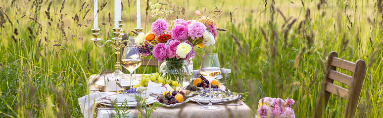 Elegant wedding or private party for a loving couple table setting outdoors in the garden. Beautiful decor with fresh ranunculi flowers, candles. Gourmet cheese board, fruits, white wine. Banner