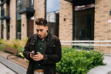 Puzzled young man standing in urban street, holding smartphone and reacting with astonishment to...