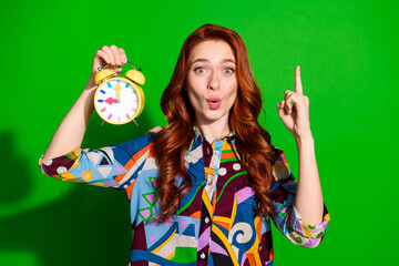 Fototapeta premium Young woman holding an alarm clock with a vibrant expression against a green background