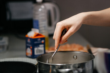 A close-up shot of a hand stirring food in a stainless steel pot on a stovetop, with a blurred background of kitchen ingredients and utensils, capturing a cozy home cooking moment
