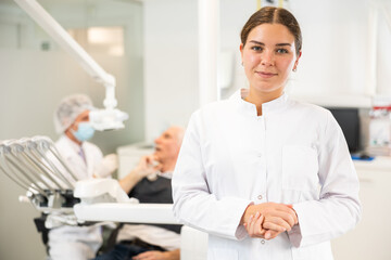 Young woman doctor is standing in dental office, clinic employee in white suit uniform near dentists workplace. Doctor enjoys working in profession.