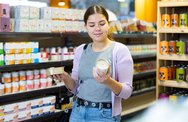 Portrait of interested young woman carefully reading labels on tin cans with choosing right hypoallergenic food for her dog in pet store