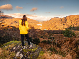 Fototapeta premium Teenager girl looking at stunning nature scenery from Ladies viewpoint in Killarney, Ireland. Travel and tourism concept. Model wearing yellow jacket and has long hair. Warm sunny day.