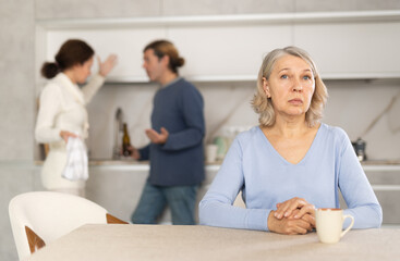 Mature woman is sitting in kitchen and is upset by quarrel between her son and daughter-in-law....