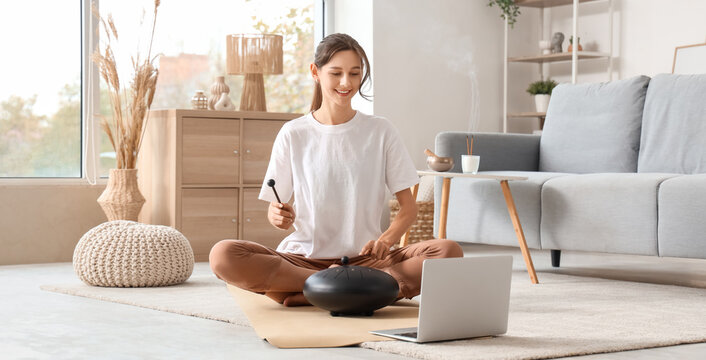 Meditating young woman with laptop playing glucophone on yoga mat at home