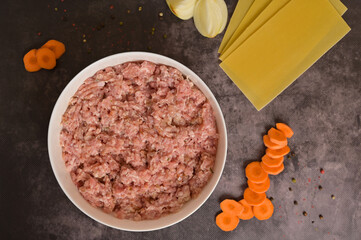 Top view raw ground minced meat in white bowl. Lasagne pasta sheets, onion, carrot, pepper. Black background. Homemade lasagna cooking preparation
