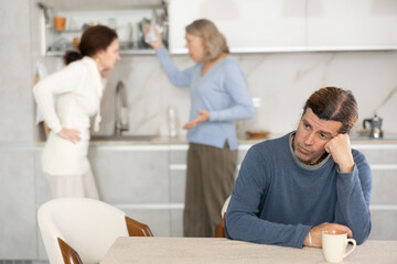 Obraz premium Man is sitting in kitchen and is upset by quarrel between his wife and elderly mother-in-law. Mature mother-in-law screams and insults middle-aged female.