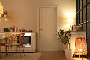 Interior of modern kitchen with glowing lamps, table, white counters and door at evening