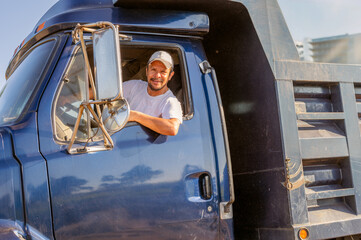 Cargo truck driver, sitting inside the truck, smiles at the camera.