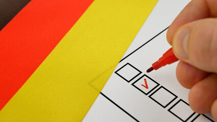 Voting in Germany. Close-up of voters hand using red marker to select candidate. Checkmark placed in checkbox. Background with German flag. Representation of political decision, fair elections