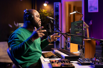 Young singer recording his new composed song on microphone, editing the tracks with mixing console and professional software. Artist manipulating sounds to create a good soundtrack.