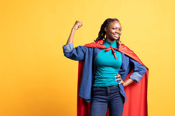 African american girl flexing arm biceps, wearing superhero costume for Halloween, isolated over studio background. Teenager dressed as comic book hero for event, showing power and strength © DC Studio