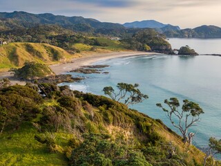 Naklejka premium Coastal New Zealand landscape. Gentle waves lap a sandy beach, framed by lush greenery and hills. Tranquil scene. Elliot Bay, Rawhiti, Northland, New Zealand