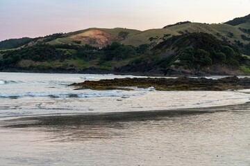 Coastal scene at dawn, with gentle waves lapping at the shore. Rolling hills and lush vegetation meet the ocean. Tranquil morning light. Elliot Bay, Rawhiti, Northland, New Zealand