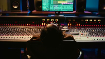 Sound designer relaxing on studio chair after mixing and mastering tracks, finishing his musical project in control room. African american audio expert producing new songs. Camera B.