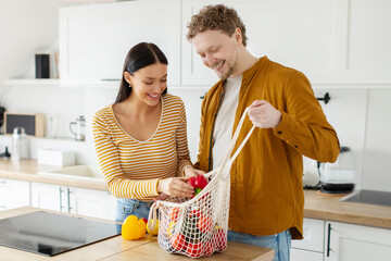 Happy spouses arriving from grocery with eco bag, unpacking fresh vegetables together in kitchen interior, copy space