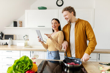 Happy couple with digital tablet cooking together and searching recipes online in kitchen at home. Cook meal application concept