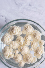 cauliflower florets soaking in a glass bowl of water. On a white marble background