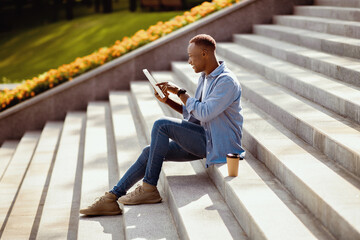 Side view of happy black guy using tablet computer on stone stairs at city park