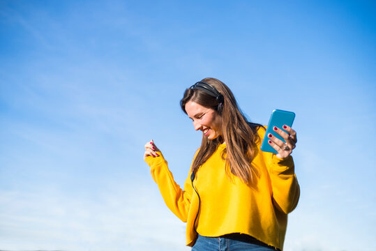Woman listening to music with headphones and smartphone, dancing under blue sky