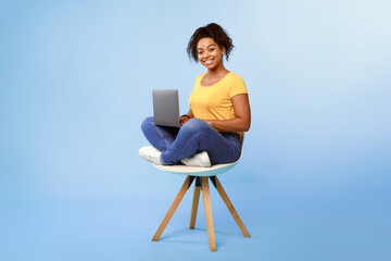 Happy young African American lady sitting on chair, using laptop, studying online or having business meeting on blue studio background. Joyful woman having remote class or watching webinar, copy space