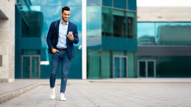 Confident arabic guy programmer with backpack walking by street, checking his mobile phone, chatting with friends, going home from office, panorama with copy space, full length