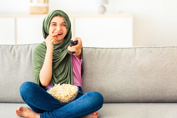 Home entertainments. Full length of Muslim teenage girl in hijab with remote control eating popcorn...