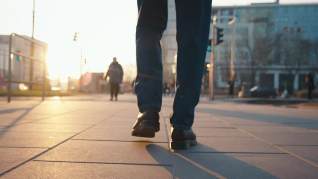 Back view following male feet steps walking pedestrian crossing street on sunny autumn day. Man wearing leather boots goes crossroad. Low angle people legs stepping backlit warm sunlight