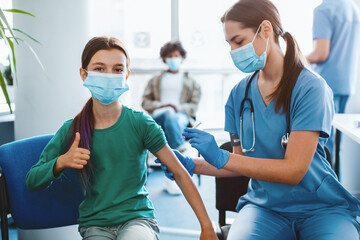 Successful flu Vaccination Concept. Positive Girl In Face Mask Gesturing Thumb Up While Getting Vaccine Injection In Shoulder, Posing Sitting At Clinic Hospital, Blurred Background. Immunization