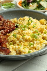 Tasty brunch. Delicious scrambled eggs, bacon and salad served on white wooden table, closeup
